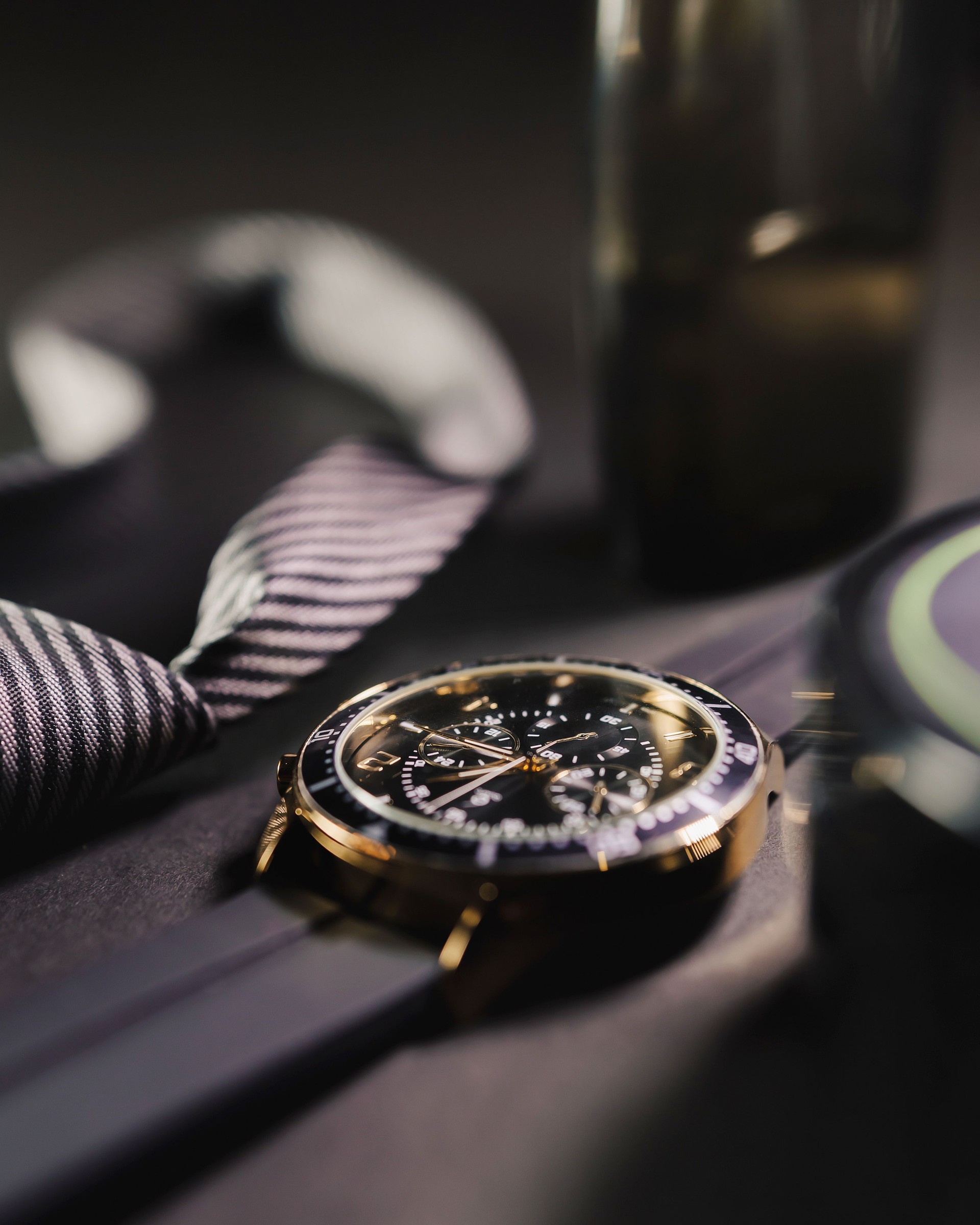 Banner of glasses, tie, and a bottle of cologne. The bottle of cologne is on a table next to a watch in an elegant setting