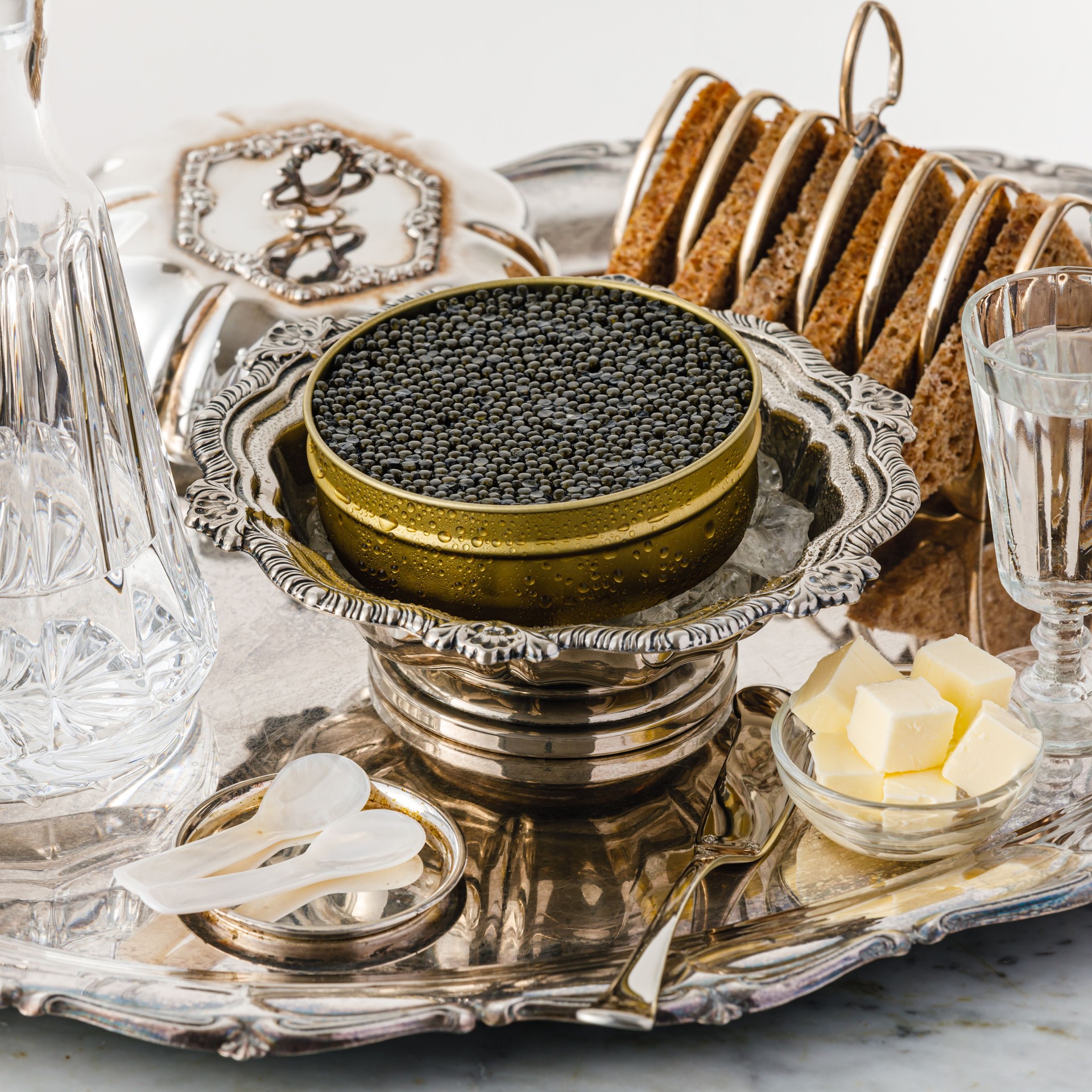 Black caviar on ice in silver bowl, Vodka and bread on white marble table close-up